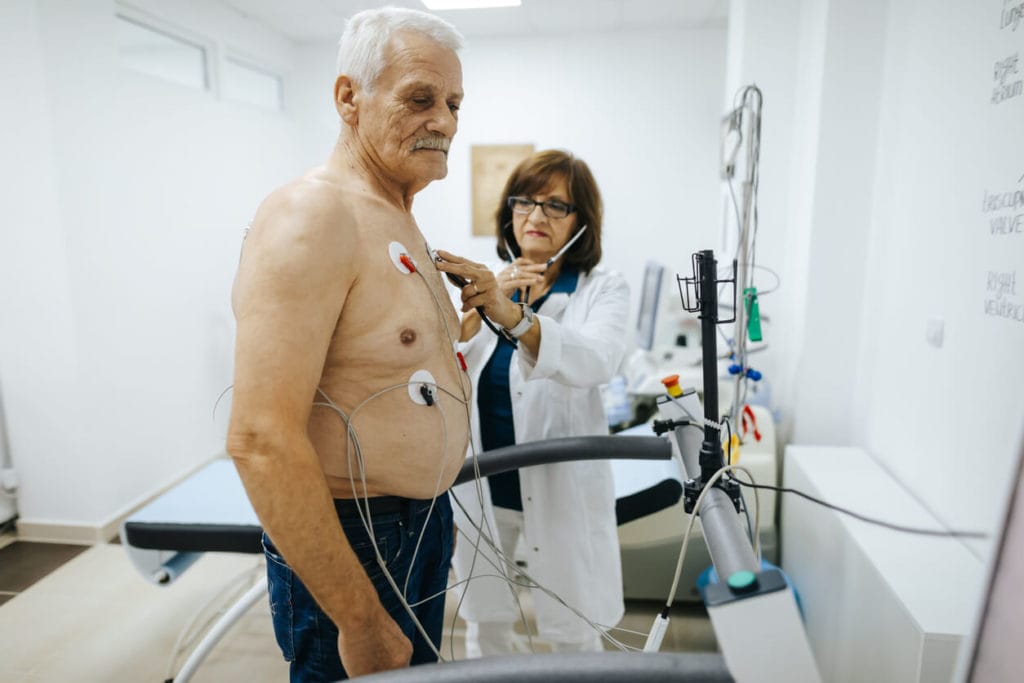 A mature female doctor examines a senior male patient during a heart stress test on a treadmill at a cardiology clinic.
