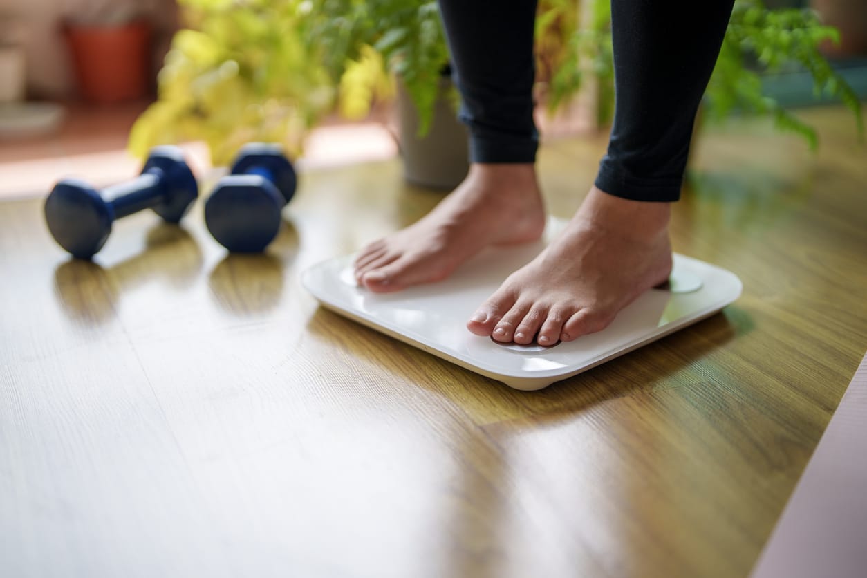 Person standing on a digital bathroom scale at home with blue dumbbells and indoor plants in the background, representing weight management