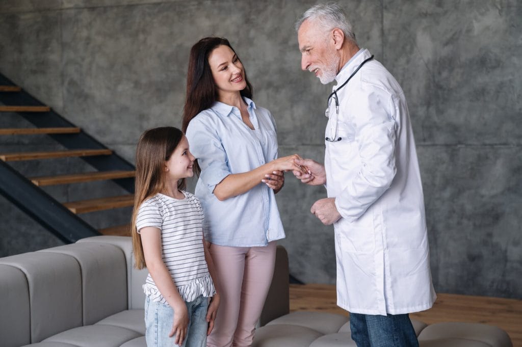 A Woman and her child are standing in the living room, greeting a senior man who is their family practice doctor.